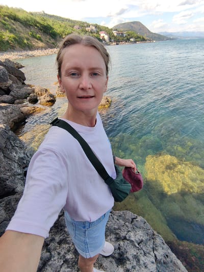 A woman with blonde hair and blue eyes is taking a selfie by the sea in Utios, Ukraine. She is wearing a light pink t-shirt, denim shorts, and white sneakers. She has a dark green bag slung across her body and is holding a maroon hat in her right hand. The woman is standing on a rocky shore, with the clear turquoise water of the sea in front of her. The background shows a coastline with green hills and buildings, and a mountain in the distance under a cloudy sky.