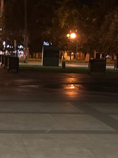 A night photograph captures a street scene in Siedlce, Poland, likely near the intersection of Piłsudskiego Street and Sienkiewicza Street, as indicated by the memorial plaque visible in the background which is commonly located in this area. The image depicts a wet asphalt road reflecting the warm glow of a street lamp with two bulbs. Adjacent to the road is a patch of green grass, and beyond that, trees with dark foliage. Several dark, rectangular objects, possibly planters or benches, are positioned along the grassy area. In the background, a tall, thin, dark pole stands prominently, possibly a lamppost or utility pole. The overall atmosphere is dimly lit, characteristic of nighttime in an urban environment.