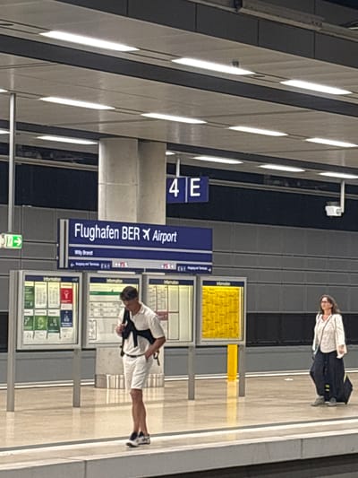 The image depicts an indoor train station platform at Berlin Brandenburg Airport (BER) in Schönefeld, Germany. A prominent blue overhead sign with a plane icon reads "Flughafen BER Airport" and "Willy Brandt," indicating the airport's name. Below this, smaller white text and symbols guide towards "Ausgang und Terminals." To the right, another blue sign displays "4 E." A green illuminated arrow sign points to the left, indicating an exit.

In the foreground, a young adult male stands on the light-colored, reflective platform, looking downwards. He wears a white polo shirt, white shorts, and black and white athletic shoes, with a black strap across his chest. His right hand is in his pocket. To the right and further back, a middle-aged woman walks towards the left, pulling a dark wheeled suitcase with her right hand. She wears glasses, a light-colored patterned top, and dark pants or a long skirt, with a dark shoulder bag strap visible.

Three large information boards on silver stands are positioned between the man and the background wall. The leftmost board shows diagrams in green, red, and blue sections. The middle board displays schematics or maps. The rightmost board features a grid of yellow squares. The background wall is covered in dark gray horizontal tile-like panels. The ceiling is composed of light panels with multiple rectangular fluorescent light fixtures. A concrete support pillar is visible behind the information boards, and a white security camera is mounted on the ceiling structure on the right side of the image.