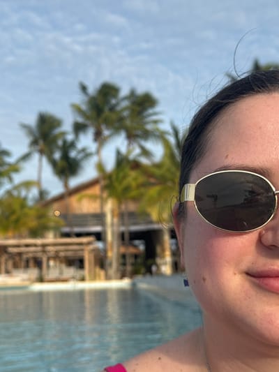 The image depicts a partial, close-up view of a person positioned on the right side of the frame, against a blurred tropical resort background in Zanzibar, Tanzania.
**Subject:** The visible individual is a female, presented from the right side of her face, neck, and upper torso. She wears gold-framed, oval sunglasses with dark, reflective lenses that obscure her right eye. Her dark hair is pulled back, with minor wisps visible near her forehead. Her skin appears natural with visible small moles or freckles on her right cheek and shoulder. A small, dark blue, rectangular earring is present on her right earlobe, and a thin silver chain is visible around her neck. A fuchsia or dark pink strap, consistent with a swimsuit or top, is visible over her left shoulder. The corner of her mouth is slightly upturned, indicating a subtle smile.
**Environment:** The background is significantly out of focus. A blue swimming pool with ripples occupies the lower-left and mid-ground. Beyond the pool, several tall palm trees are visible. A large, multi-story building, characteristic of a resort or lodge, is present in the mid-background, featuring wooden construction and a prominent roof structure. To its left, a smaller, open-air wooden structure with a pergola-like roof is partially visible. The sky above is light blue with scattered white clouds. A distinct orange-yellow circular light flare or bokeh effect is visible in the upper-left background, near the main building. The scene is illuminated by natural daylight.