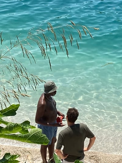 Two men stand on a pebble beach next to crystal-clear turquoise water. The man on the left, wearing a green bucket hat and blue shorts, is darker-skinned and appears to be talking to the man on the right. He is holding a red object in his right hand. The man on the right, with curly brown hair, is wearing a dark green t-shirt and light-colored shorts. He has his hands on his hips and is looking out at the water. In the foreground, lush green leaves and dry grass stalks frame the scene. The water is shallow and transparent, revealing the pebble seabed. The location appears to be a scenic coastal area, consistent with Perast, Montenegro, known for its clear waters and picturesque beaches.