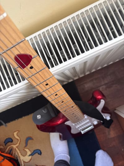 An overhead shot captures an indoor scene in Glava, Bulgaria, featuring a red electric guitar and a white radiator. The guitar's light wooden fretboard, adorned with silver strings and black dot inlays, extends diagonally from the bottom center, with a red guitar pick tucked between the strings near the first fret. The body of the electric guitar, a glossy red with a silver-colored pickguard and bridge, rests on a dark wooden floor. A black strap, featuring a small white stylized "C" or crescent logo, is attached to the guitar. To the left, a white radiator with horizontal slats, appearing somewhat dusty, runs parallel to the guitar's fretboard. In the background, a beige rug with blue and light brown patterns is visible, alongside an orange object, possibly a bag or backpack. The feet of a person, wearing white socks, are partially visible on the rug and wooden floor in the upper right and lower right corners. The composition suggests a casual, domestic setting, possibly a practice space or living room.