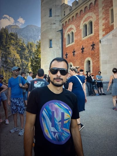 The image shows a group of people in front of a castle-like building. In the foreground, a man in a black t-shirt with a colorful graphic is taking a selfie. Behind him, other people are visible, some looking at the camera, others engaged in conversation. The building appears to be a historical structure with red brick and stone accents, featuring arched windows. The background reveals a mountainous landscape with trees. The scene is outdoors, with a paved walkway in front of the building. The time of day appears to be daytime, with natural lighting. The location is identified as the city of Kozlovets, Bulgaria.