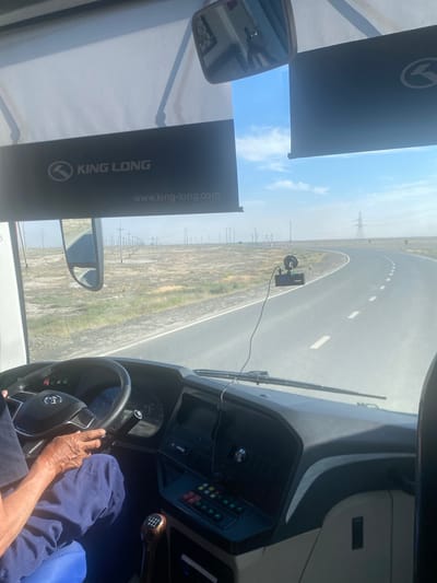 The image provides a driver's perspective from inside a King Long bus, traveling on a paved road. The bus is moving through a dry, semi-arid landscape characterized by sparse vegetation, dirt tracks, and numerous utility poles stretching along the roadside, consistent with the environment of Қосшағыл ауылдық округі, Kazakhstan. The driver, an elderly individual with weathered hands, is visible in the lower left, gripping the steering wheel and wearing blue trousers. The dashboard is equipped with a digital display, buttons, and a manual gear shifter. A dashcam is mounted on the windshield. The sky is clear and bright blue, with some wispy clouds. The road curves ahead, disappearing into the distance under the bright sunlight.
