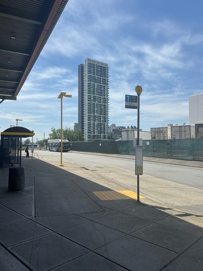 This is a landscape-oriented photograph taken outdoors, likely in the late morning or early afternoon given the harsh shadows cast by the sun. The scene depicts a bus stop area.
In the foreground, a dark, corrugated metal structure is visible on the left, possibly part of a building or shelter. The ground is paved with large, gray concrete tiles, with a textured yellow tactile paving strip running parallel to the walkway. A bus stop sign is prominent, featuring a yellow circle with the number "3" inside, and a blue "T" logo. Below this, text indicates "Bus Stop," a route number "58083," and destinations "159 Coquitlam Central Station." A smaller sign with a QR code is also attached to the pole.
In the background, several elements contribute to the urban setting. A modern, tall residential building with a grid-like facade dominates the skyline. Further back, shipping containers are stacked and secured by a chain-link fence, with company names like "J.B HUNT" and "H.O.WOLDING" visible on them. A public bus is stopped at the bus stop. Trees and other smaller buildings are also visible in the distance.
The sky is a bright blue with scattered wispy white clouds. The overall impression is of a functional, urban transit hub on a clear, sunny day. No people are clearly discernible in the image, but the presence of a bus stop and bus suggests a setting where people would be present. The location is identified as New Westminster, Canada.
