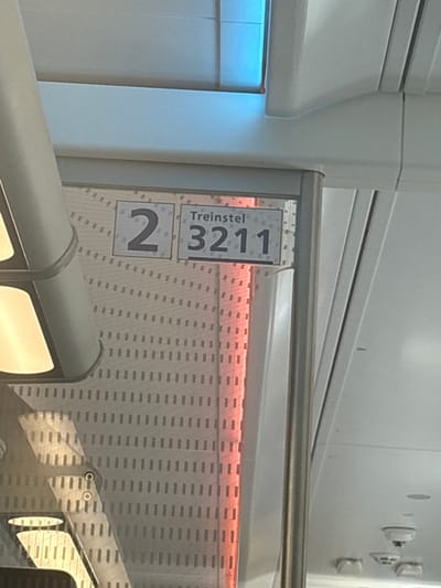 This image shows an interior view of what appears to be a modern train or public transport vehicle. The main subjects are the signs indicating "Treinstel 2 3211". The sign "Treinstel" is written in a dark font on a white background. Below it, the number "2" is prominently displayed in a large square box, also in dark font on a white background. To the right of the "2", another sign shows the number "3211" in a larger font, with "3" and "2" above the "11". The signs are mounted on a railing that runs horizontally across the image.
Behind the railing, there is a textured wall with a repeating pattern of small dark rectangles against a lighter background. A band of warm red light illuminates the upper part of this wall. The overall setting is bright and clean, suggesting a well-maintained interior. To the left of the signs, a smooth, light-colored surface with a bluish glow is visible, possibly a window or part of the vehicle's interior design.
In the foreground, a rounded, light-colored handrail is present, with another handrail above it. Reflections of illuminated elements are visible on the lower handrail, suggesting there are lights within the vehicle. The image seems to be taken during the day, given the overall brightness, though the internal lighting, particularly the red band, creates a distinct ambiance. There are no people visible in the frame. The text "Treinstel 2 3211" clearly indicates the train set number. The location context of Eindhoven, Netherlands, might suggest this is a Dutch train.