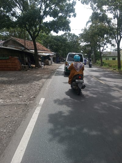 The image captures a daytime street scene in Chhenangri_Rishor, Bhutan, from a rear perspective behind a moving dark-colored scooter. An individual wearing a light blue helmet, an orange-brown garment, and a patterned head covering operates the scooter, which displays license plate "F 6894 ZD". Directly ahead, a white minivan travels in the same direction. Further down the asphalt road, a second motorcycle and a blue/purple vehicle are visible. The road features a solid white edge line on the left, bordering an unpaved dirt and gravel shoulder. To the left, a row of low-rise structures includes a building with a red tiled roof and another with a corrugated metal roof and slatted siding. Stacks of reddish-brown tiles are present outside the leftmost building. Large trees with green foliage line both sides of the road, casting shadows on the asphalt. On the right side, a grassy field precedes a light green/blue single-story house and a distant utility pole. The sky overhead is bright.