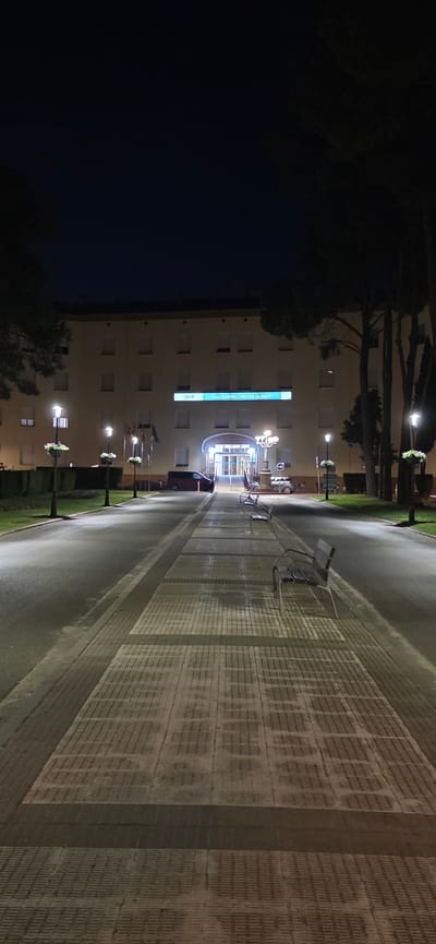 A night time, eye-level, vertical, full shot shows the exterior entrance of a hospital at the end of a pathway. The building is light beige with multiple rows of square windows. Above the entrance, a lit blue sign reads "SALUD SAGRADO CORAZÓN DE JESÚS". The entrance itself is an archway with glass doors. Flanking the path are lampposts with hanging flower baskets. Three benches are visible along the pathway. To the right, trees form a dark silhouette against the night sky. The pathway is paved with light-colored rectangular tiles with dark grout, and the sides of the path are lined with dark gray concrete. A red van is parked to the left of the entrance, and a silver car is parked to the right. The scene is in Huesca, Spain.