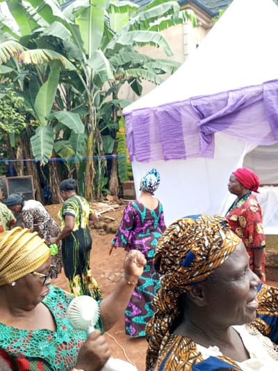 The image displays an outdoor gathering in Oko Town, Nigeria, featuring an unpaved reddish-brown earth ground and dense tropical foliage, predominantly large banana trees, in the background. A white event tent with purple draped fabric along its valance is situated on the right side, partially obscuring a light-colored building with a dark roofline in the upper right background. A wooden frame structure is partially visible in the far left background, near the banana trees.
At least seven adult females are present, all dressed in patterned traditional Nigerian garments, including matching headwraps (geles). In the foreground on the left, an older woman with dark skin, wearing glasses, a green patterned top, and a gold gele, holds a small white portable fan in her right hand, directing it towards herself. Her left arm is slightly raised, revealing a gold-colored bracelet. To her immediate right, another older woman with dark skin wears a brown, gold, and blue patterned gele and top, viewed in profile, looking towards the right with a neutral expression.
In the midground, a woman in a dark patterned dress with green and yellow accents is seen walking away from the camera. Beside her, another woman in a patterned dress is bent over, possibly attending to something on the ground. Further right, a woman in a purple, brown, and teal patterned long-sleeved dress and matching headwrap faces away from the camera, moving towards the tent. Near the tent on the right, an older woman in a red headwrap and red and gold patterned top stands, looking towards the right. Piles of reddish-brown material are visible on the ground in the mid-left background.
