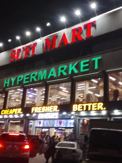The scene depicts the exterior of the Sufi Mart Hypermarket at night in Babban Giji, Nigeria. The multi-story commercial building is brightly illuminated against a dark sky. At the top, a large sign displays "SUFI MART" in red illuminated capital letters. Below this, "HYPERMARKET" is visible in prominent green illuminated capital letters. The mid-section of the building features multiple large, glass-paneled windows, revealing brightly lit interior retail spaces. Directly below these windows, three distinct yellow illuminated signs read "CHEAPER.", "FRESHER.", and "BETTER." from left to right.

At street level, below these yellow signs, an active entrance is visible. An illuminated sign above the entrance reads "ENTRANCE," and another sign below it displays "I ❤️ KANO I ❤️ Biffi na". A number of individuals are present on the sidewalk and congregating near the entrance, indicating pedestrian traffic. In the foreground, a street or parking area contains several vehicles. A white car with illuminated red taillights is positioned on the left, facing away from the viewer. A lighter-colored sedan is partially visible behind it. On the far right, a dark-colored van or SUV is parked or moving. The dominant lighting originates from the building's extensive signage and interior lights, with additional streetlights visible overhead.