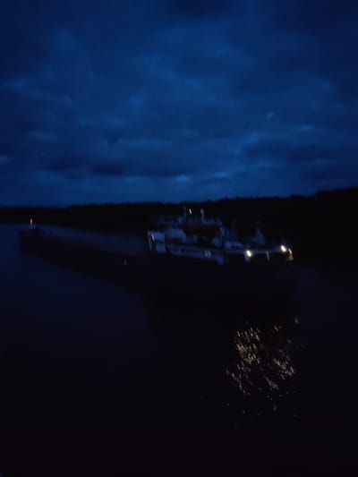 A nighttime scene shows a large barge on a dark body of water. The barge has several white structures on it, lit by a few bright lights. Dark trees line the horizon. The sky is filled with dark, cloudy formations. Reflections of the lights on the barge are visible in the water. The location is not specified. The exact city location is not discernable from the image.