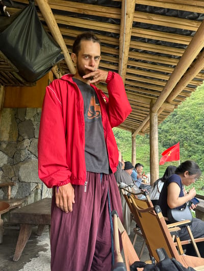 Captured in Yên Minh Commune, Vietnam, the photograph presents a man in a red jacket and maroon pants, positioned in a structure with bamboo-like supports and wooden crossbeams. The setting suggests an outdoor or semi-enclosed space, possibly a shelter or seating area. He is seen in the center. There is a Vietnamese flag in the background, along with other people sitting and using mobile devices. A black trash bag is nearby and the structure seems to be built from wood. The wall is made of stacked stones.