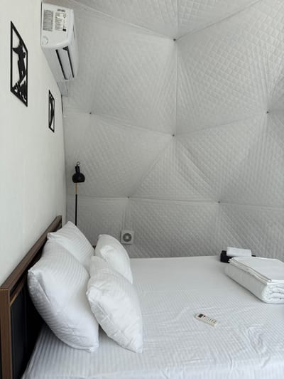 The image shows an interior view of a geodesic dome room in Nechkino, Russia, furnished as a bedroom. The dominant color scheme is white, with the geodesic walls featuring a quilted texture. A bed with white striped bedding and several white pillows is positioned in the foreground. A dark wooden headboard is visible on the left.

On the wall to the left, an air conditioning unit is mounted near the ceiling. Below it are two pieces of black geometric wall art. A black floor lamp with a round head stands near the head of the bed. On the quilted wall behind the bed, a small white ventilation grate is visible.

To the right side of the bed, a stack of folded white towels and what appears to be dark towels or bedding is placed. A white remote control rests on the bedsheets. No people are present in the image. The scene depicts a tidy and minimalist bedroom environment.
