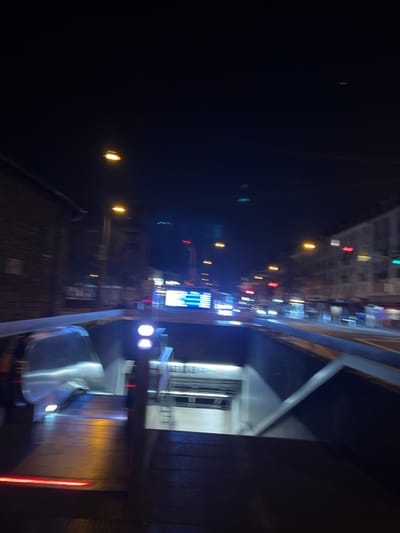 The image shows a view from a moving vehicle on a bridge in Köln, Germany (latitude: 50.939, longitude: 7.009). The bridge is made of metal and has a reflective surface. There are multiple streetlights illuminating the road below.  A few cars are visible in the distance. The image is taken at night, and the city lights and reflections on the water create a blurry, impressionistic effect. The camera is pointing downwards toward the ground.  The image captures a moment of urban movement.