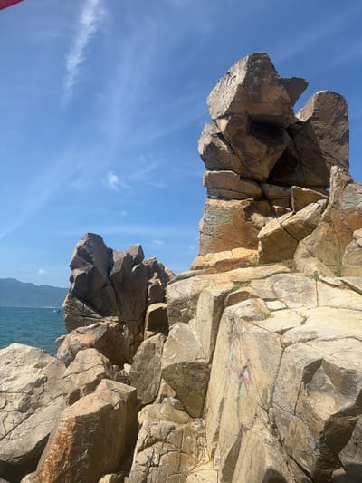This image captures a section of the coastline in Vĩnh Lương, Vietnam, characterized by large, weathered rock formations against a clear blue sky with scattered white clouds. The rocks are a mix of tan, brown, and grey, with visible cracks and fissures indicating geological activity and erosion. Some rocks are stacked precariously, creating a rugged and dramatic landscape. Green vegetation sprouts from some of the crevices in the rocks. In the background, the turquoise ocean stretches towards a hazy mountain range. A small, dark-hulled boat is visible on the water to the left of the frame. The lighting suggests it is daytime, with sunlight casting shadows and highlighting the textures of the rock surfaces.