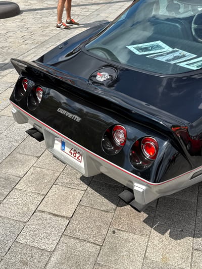 The image shows the rear of a black Corvette Stingray parked on a cobblestone street in Bruges, Belgium. The car has a silver trim along the rear bumper and fender, with red pinstriping. Prominently displayed on the rear is a license plate with the number "482" and the Belgian flag emblem. The car's taillights are circular and red, and it has dual exhaust pipes visible.
Reflected in the car's window are what appear to be stickers or prints of older vehicles. To the left of the car, a person's legs and feet are visible, wearing orange and white sneakers, suggesting they are either the owner or an onlooker. The lighting indicates it is daytime, likely sunny given the clear reflections. The scene is set on a well-maintained cobblestone street, typical of historic European cities like Bruges. The overall impression is that of a car show or a gathering of classic car enthusiasts. The image is oriented vertically.