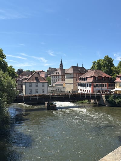 This is a landscape photograph of the Regnitz River in Bamberg, Germany, taken on a sunny day. The river flows from the foreground towards the background, with a bridge spanning across it. Along the riverbanks, there are several buildings with red tiled roofs, typical of Bamberg's historic architecture. Some of these buildings appear to be restaurants or cafes, with outdoor seating visible on the bridge and along the bank. Trees line the banks of the river, with lush green foliage. The sky is blue with a few wispy clouds. The scene depicts a serene and picturesque urban environment.