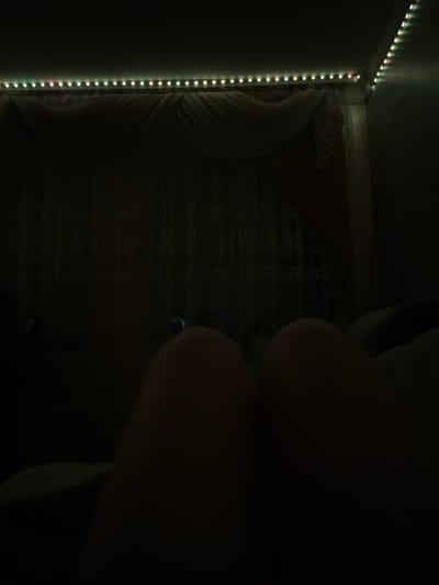 The image depicts a dimly lit room, likely a bedroom, with string lights illuminating the ceiling and upper portion of the walls. The lights appear to be LED strip lights, displaying a multicolor effect, predominantly white and green with hints of pink. In the foreground, a person's legs are visible, suggesting they are lying down or reclining. The legs are in shadow, with only their outlines discernible. Behind the legs, the background is obscured by darkness, but the faint shapes of curtains and possibly furniture can be inferred. The overall atmosphere is one of low light and intimate setting, suggestive of nighttime or a deliberately darkened space. The location specified, Slobozia-Dușca, Moldova, provides a geographical context to the scene, but the image itself does not contain any specific visual cues to confirm this location.