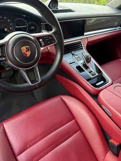 The image shows the interior of a Porsche Panamera 4. The interior features red leather seats, a black leather-wrapped steering wheel with the Porsche emblem in the center, and a glossy black center console. The dashboard shows the time as 17:06 on Saturday, May 2nd, 2026. The location is not identifiable from the image.