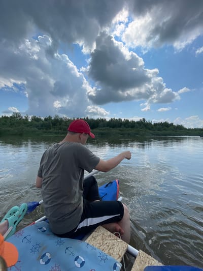 A person wearing a red cap and a grey t-shirt is seated in a raft on a body of water. The person is facing away from the camera, and their right arm is extended, with their hand appearing to be in the water. The raft has a blue and red bag and a blue mat with anchor and life preserver patterns. Light blue Crocs are visible on the raft. The water is calm with ripples. In the background, a line of dense green trees runs along the horizon. The sky is a bright blue with scattered, dramatic clouds, some dark and some white and fluffy. The scene suggests an outdoor recreational activity on a river or lake under a partly cloudy sky. The image appears to have been taken from the perspective of someone also in the raft. The general location is described as the city of Нежинка, Russia.