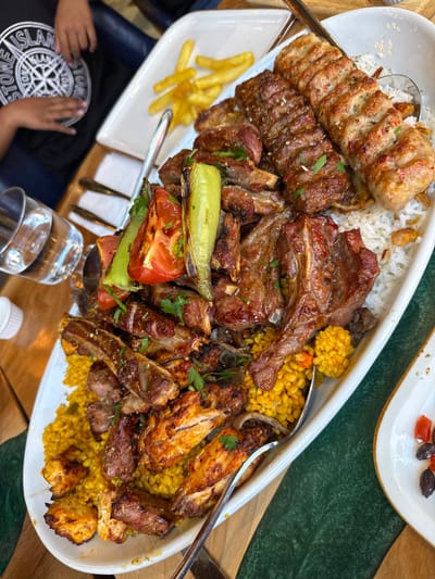 The image shows a large platter of mixed grilled meats, including kofta kebab, lamb chops, chicken wings, and diced chicken pieces, served with white rice and a yellow rice pilaf. Grilled tomato and green peppers garnish the dish, which also contains fries. Silver serving spoons are present. The platter sits on a wooden table, with a glass and a small bottle nearby. A person wearing a Stone Island T-shirt is visible on the left side of the image. Another plate containing olives and red fruits is slightly visible at the top right of the image. Without additional information or access to geolocation services, the location within Nottingham, United Kingdom cannot be ascertained.