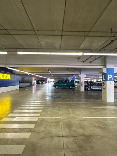 The image depicts the interior of a multi-level parking garage at IKEA in Alfafar, Spain. The concrete ceiling, supported by columns, features exposed conduits and fluorescent lighting. The floor is marked with white lines indicating parking spaces and a pedestrian crossing. In the foreground, a pedestrian crossing is visible with white stripes. To the left, a section of the IKEA store's blue facade with yellow lettering is present. In the middle ground, a teal van is parked. Further back, several cars are parked in designated spots, and shopping carts are lined up near the store entrance. A blue and white sign with "L P -2" is visible on a column, indicating parking level. The environment is illuminated by artificial lighting, creating a utilitarian atmosphere. There are no people visible in the immediate scene.