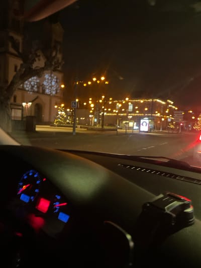 Nighttime scene viewed from inside a car. Lights illuminate a town with storefronts displaying holiday decorations. In the foreground, the car's dashboard is partially visible with illuminated gauges and indicators. The car's interior is dark. A phone mount is visible on the dashboard. The reflections on the car's windshield add to the light in the frame, obscuring the view.