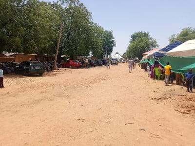 The image shows a dirt road lined with parked cars and motorcycles on the left, and a row of tents on the right. There are several people walking along the road, some of them are sitting inside the tents, some are standing, and one person is walking toward the tents. The environment is mostly dirt and dry grass, with some trees in the background. This scene is located in Zum, Nigeria, at coordinates 10.116 latitude and 13.046 longitude.