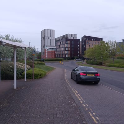 The image captures an urban or suburban street scene, likely in Coventry, United Kingdom. The perspective is from a pedestrian area, looking along a paved path and a road towards several modern buildings under an overcast sky.
In the foreground, on the left, is part of a bus stop shelter with a light-colored, possibly metallic or solar-paneled roof, supported by white poles. Beside it, a red pole holds a standard UK bus stop sign featuring a black bus icon and the text "Bus Stop". Below this, a smaller yellow sign with partially legible text, possibly "All routes here", is attached. The ground is covered with dark grey block paving, resembling cobblestones, with patches of moss or debris. Lush green bushes and some white-flowering plants line the path.
A dark grey car, appearing to be a BMW 3-series, is seen from the rear on the asphalt road, positioned as if it's about to turn or proceed. Its red taillights are visible, and the yellow UK license plate "Y44 KBY" is clearly readable. Double yellow lines, indicating parking restrictions, are painted along the curb separating the road from the pedestrian path. Further down the road, two yellow bollards and a blue "keep left" arrow sign for a mini-roundabout or junction are visible.
The background is dominated by several contemporary buildings. To the left, a tall, multi-story building features a facade with vertical silver and white striped panels. To its right, a larger complex combines dark brown sections with lighter, possibly brick-toned or terracotta, panels and numerous windows. Another building with similar modern architecture is partially visible further right, nestled amongst trees.
The scene depicts an overcast daytime, with soft, diffused light and no direct sunlight. No people are visible, and the overall atmosphere is quiet and functional.