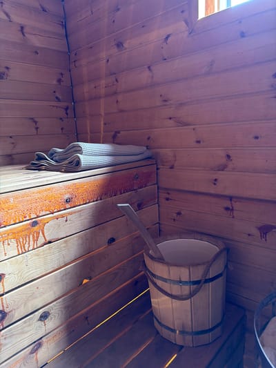 The image shows the interior of a wooden sauna. A corner of the room is visible, with walls and benches made of light-colored wood planks. Two folded towels, one lighter and one darker, are placed on the upper bench. To the right, a wooden bucket with a rope handle is positioned on a lower bench. A wooden ladle rests inside the bucket. The wood planks on the lower benches show dark, dripping stains, possibly from sap or resin. The scene is lit by natural light coming from an unseen window or opening at the top right of the frame. There are no people present in the image. The overall impression is one of a rustic and functional sauna environment.