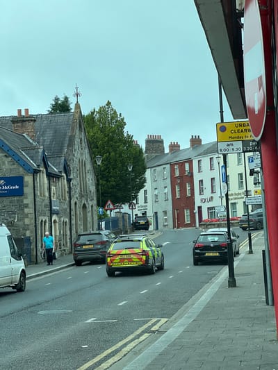 The image depicts a street scene in Omagh, United Kingdom, on a cloudy day. The perspective is from a vehicle, looking forward. The street is paved, with double yellow lines marking the edges of lanes and a pedestrian crossing visible in the distance. On the left side of the street, a white van is parked. Further along, a building with a stone facade and a dark grey roof is visible, with a blue sign that reads "McGrade Solicitors." A man in a light blue t-shirt and dark trousers, carrying a bag and a white cane, walks along the pavement.
To the right of this building, a grey SUV with license plate XIC 5879 is parked. Further down the street, a police car with reflective yellow and green markings and license plate RGZ 6916 is driving. Next to it, a black Volkswagen Golf with license plate UHZ 6808 is also in motion. The right side of the street features a row of terraced houses painted in various colors, including white, red, and cream. A sign for "FOX LAW" is visible on one of these buildings. A pedestrian walkway runs alongside these houses, lined with lampposts. Traffic signs are present, including a "No Entry" sign and parking restriction signs. The overall environment appears to be a commercial or residential area of a town or city.