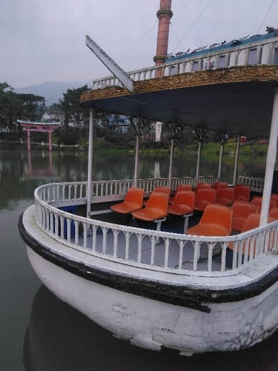 The image depicts a stationary, multi-tiered decorative boat moored in a body of still, dark water under an overcast sky. No individuals are visible.
The primary subject, the boat, features a white hull exhibiting surface degradation including chipped paint and staining, with a black protective strip lining the waterline. Its lower deck is enclosed by white baluster-style railings. Inside, a minimum of twelve unoccupied bright orange plastic seats are arranged in multiple rows. An overhead blue canopy is supported by white vertical posts. Above the canopy, an ornate gold-colored trim with intricate scrollwork details the edge of the upper structure. A prominent, tall, reddish-brown cylindrical structure, consistent with a chimney or mast, extends vertically from the boat's upper tier. The boat's pointed bow is visible in the lower left, partially submerged.
The background across the water displays a shoreline densely covered with green trees and foliage. On the opposing bank, a distinct pink Japanese torii gate, featuring a dark roof, is partially visible amidst the greenery. Additional buildings, including a white structure with dark roofs, are interspersed among the trees. Distant mountainous terrain is discernible under a uniformly gray, overcast sky.
This scene is located at the lake in Parque La Sabana, Ciudad de San José, Costa Rica, characterized by the presence of the decorative boat (known as the "USS San José" replica) and the nearby Japanese torii gate, which is associated with Parque Okayama adjacent to La Sabana.