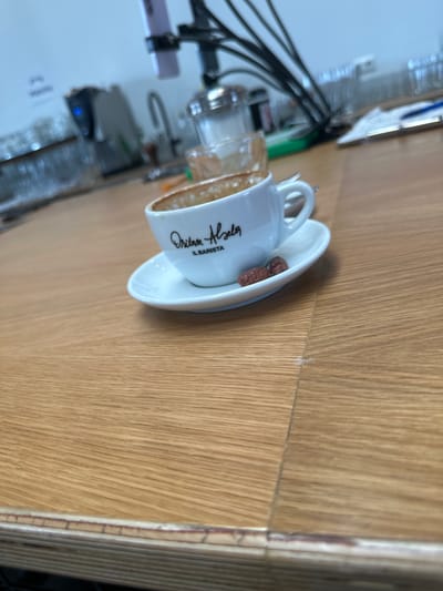 The image displays an espresso cup and saucer resting on a light brown wooden countertop within the Oritam Helsa IL BARISTA establishment, located at Kanalstraße 15, 40667 Meerbusch, Germany. The white ceramic espresso cup, bearing the black script "Oritam Helsa IL BARISTA," is positioned centrally, slightly to the right, and contains visible brown coffee residue, indicating recent consumption. A small silver spoon is situated on the saucer behind the cup. Two small, reddish-brown, coated items (likely sugared nuts) are present on the saucer next to the cup. The wooden surface exhibits a pronounced grain pattern and a visible seam line between two planks. The background, largely out of focus, reveals elements of a commercial setting: a dark appliance with a blue illuminated indicator light, stacks of clear glassware, a silver-toned dispenser, a flexible mounting arm holding a light purple smartphone, and a white sheet of paper with a blue pen. No individuals are present in the frame. The scene is brightly and evenly lit.