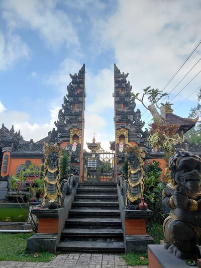The image depicts the entrance to a Balinese temple in Sukasada, Indonesia, under a partly cloudy sky. The foreground is dominated by a set of stone stairs leading up to an ornate gateway. Flanking the stairs are two large stone statues of guardian figures, one on the left appearing serpentine and the other on the right resembling a demon. These statues are adorned with gold detailing. The gateway itself is an imposing structure with intricate carvings and decorative elements, featuring orange brickwork and darker stone accents. Beyond the gateway, a glimpse of the temple's architecture is visible, including a tiled roof and more decorative stonework. Lush green vegetation surrounds the entrance, including potted plants and trees. A small pond or water feature is visible to the left of the stairs, with what appear to be koi fish swimming within. Power lines are strung across the upper right portion of the image. The overall scene conveys a sense of traditional Balinese artistry and spiritual architecture.