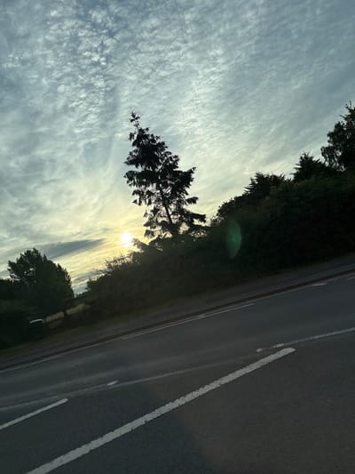 The image depicts a slightly tilted, outdoor scene of a road, trees, and sky, likely taken in the city of South Staffordshire, United Kingdom. The road, paved with asphalt, is visible in the foreground and extends diagonally into the background. White lane markings are present on the road. Flanking the road is a dense line of trees and foliage, silhouetted against a bright, hazy sky. A prominent evergreen tree, its branches silhouetted, stands out against the sky. The sun is visible in the upper left quadrant of the image, partially obscured by clouds, casting a soft, yellow light. The sky is filled with scattered, wispy clouds, creating a textured pattern. A faint green lens flare is visible on the right side of the image, near the trees. The overall impression is one of a tranquil, possibly early morning or late afternoon, rural or semi-rural road setting. There are no people or vehicles visible in the frame.