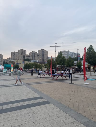 The image depicts a public outdoor space in Zelenograd, Russia, characterized by a paved plaza and surrounding buildings. In the foreground, a man in a grey t-shirt and shorts is walking across the plaza, facing left. Two ping pong tables are set up on the plaza, with several people engaged in playing or watching. One table has two individuals playing, one of whom is in motion. Another table has a group of people standing around it.
Red banners are visible on flagpoles in the background, and trees are planted along the plaza's edge. Behind the plaza, multi-story residential buildings with light-colored facades dominate the skyline. The sky is overcast.
The area appears to be a recreational or gathering space within the city, possibly a park or a pedestrian zone, given the presence of recreational equipment and the public setting. The architecture of the buildings suggests a Soviet-era or post-Soviet urban environment. The overall atmosphere seems casual and relaxed, with people enjoying outdoor activities.