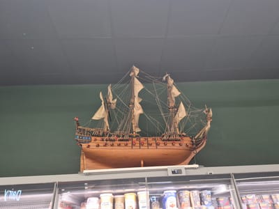 An overhead shot captures a detailed wooden model of a galleon ship mounted against a dark green wall. The ship features multiple masts, sails, rigging, and cannons along its hull. Below the ship, a commercial refrigeration unit is visible, displaying various yogurt and ice cream containers with labels such as "Glenisk Organic," "Superior Organic," and "Cluster." The unit has a digital display showing "30". The lighting suggests an indoor environment, likely a supermarket or store. The text "kriss" is faintly visible on the glass of the refrigeration unit. The location is the SuperValu supermarket in Clonakilty, Ireland.