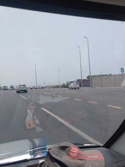 The image shows a view from inside a vehicle, likely a car or truck, driving on a highway. The road is paved and marked with yellow and white lines. Several other vehicles are visible on the road, including a white pickup truck, a silver sedan, and several other cars in the distance. The sky is overcast, giving the scene a somewhat muted appearance.
In the foreground, part of the vehicle's dashboard is visible, with a baseball cap resting on it. The reflection of a gas station pump appears on the windshield.
In the background, there is a concrete wall, light poles, and some greenery. Based on visual characteristics such as the light poles and infrastructure, this location is likely in or near Jeddah, Saudi Arabia.