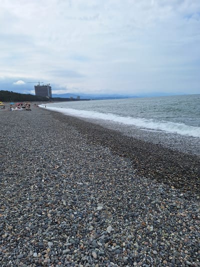 The image depicts a pebble beach in Kobuleti, Georgia, under a cloudy sky. The foreground is dominated by a vast expanse of smooth, dark pebbles. In the middle ground, a line of people are visible, some reclining on lounge chairs and others walking along the shore. The sea, with gentle waves breaking on the beach, extends to the horizon. In the background, a large, unfinished hotel building with a construction crane is prominent, alongside other buildings and a treeline. Mountains are faintly visible in the distance. The overall scene suggests a moderately populated beach with ongoing development.