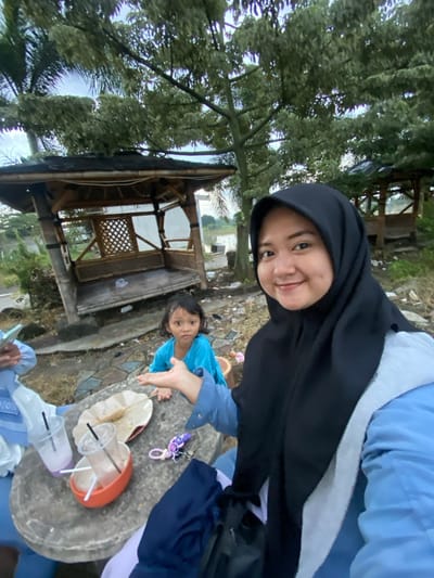 The image depicts an outdoor scene in Kalitengah, Indonesia. In the immediate foreground, a woman in a black hijab and a light blue long-sleeved top is taking a selfie, smiling directly at the camera. Her right hand is visible, positioned near a round, gray stone or concrete table. On the table, there are two empty clear plastic cups with straws, an orange bowl containing some residue, a crumpled paper wrapper with a wooden skewer, and a purple keychain with a white attachment.

To the left of the table, another person's arm and hand are visible, holding a smartphone, also wearing a light blue sleeve.

Further back, a young child wearing a bright blue short-sleeved top is looking towards the camera with a neutral expression, positioned behind the table with both hands slightly open and extended forward.

The background features several trees with light green foliage under an overcast sky. Two rustic bamboo gazebos with dark roofs are visible. The gazebo on the left is more prominent, appearing weathered with some structural elements exposed. Beyond the gazebos and trees, a body of water is discernible, possibly a river or lake. The ground is uneven, composed of dirt and scattered debris.