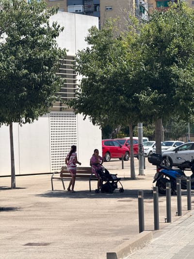 The image shows an outdoor scene in l'Hospitalet de Llobregat, Spain, on a sunny day. Two women are seated on a bench in the foreground, with a baby stroller next to one of them. One woman wears a pink patterned outfit, and the other wears a white and red patterned outfit. Behind the bench, a modern white building with a textured facade and horizontal window elements is visible. Several mature trees with lush green foliage are scattered throughout the scene, providing shade. In the background, cars are parked, including a red SUV, a white sedan, and a silver SUV. A blue scooter is parked on the right side of the image. Several dark cylindrical bollards are placed along the sidewalk. The ground is paved with concrete and some cobblestones.