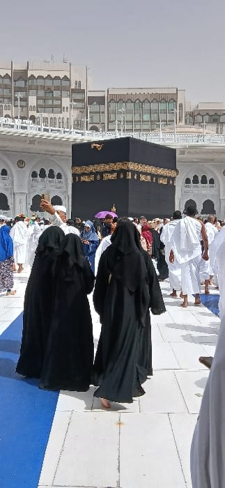 The image depicts an outdoor scene at the Grand Mosque (Masjid al-Haram) in Makkah Al Mukarramah, Saudi Arabia, with the Kaaba centrally positioned in the mid-background. The Kaaba is a prominent black cuboid structure featuring gold calligraphy and ornamentation along its upper band.
In the immediate foreground, three women, viewed from behind, walk forward. They are dressed in black abayas and head coverings; the woman on the right is barefoot. To their left, a man wearing white ihram attire and a white cap is seen holding a smartphone aloft, potentially capturing an image or video. Surrounding these individuals, a multitude of other people, primarily pilgrims, move across the scene. Many men are dressed in white ihram garments, while women wear various forms of modest attire, including black abayas and blue head coverings. One individual holds a purple umbrella open for shade.
The ground is paved with light-colored, square tiles, with a section of blue matting present along the left side of the foreground. The Kaaba is framed by the white, arched architecture and balustrades of the mosque complex. In the distant background, multi-story modern buildings with numerous windows are visible beneath a light blue, hazy sky. The environment is brightly lit, with visible shadows indicating direct sunlight.