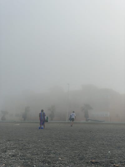 A vertical shot captures a hazy beach scene in La Línea de la Concepción, Spain. The image is dominated by a thick fog, obscuring the background and creating a soft, diffused light. Two figures are visible on a pebble-covered beach. In the mid-ground, a person wearing a white t-shirt and dark shorts is captured mid-stride, appearing to be walking or playing. Further in the foreground, another individual is seen from behind, wrapped in a colorful, striped blanket or garment in shades of blue, red, yellow, and green, with blue footwear visible. A building, possibly an apartment complex, is faintly visible through the fog in the upper left portion of the image, along with the silhouette of palm trees. A lamppost stands near the person in the mid-ground. The presence of fog suggests a morning or evening time, or perhaps a coastal weather phenomenon. The activity appears to be casual, with the individuals enjoying the beach despite the limited visibility. No text is visible in the image.