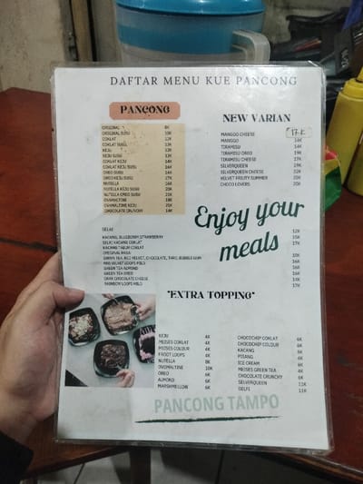 A hand holds up a laminated menu for "Kue Pancong" (Pancong Cake), likely in an indoor eatery in Dramaga, Indonesia. The menu, titled "DAFTAR MENU KUE PANCONG," details various offerings and prices, indicated in thousands of Rupiah ("K").
The main "PANCONG" section lists flavors such as Original, Susu, Coklat, Keju, Oreo, Nutella, Ovomaltine, and Chocolate Crunchy, ranging from 8K to 21K. A "NEW VARIAN" section features options like Manggo Cheese, Tiramisu, Silverqueen, Velvet Fruity Summer, and Choco Lovers, priced between 14K and 22K.
Additional categories include "SELAI" (Jams) with Kacang, Blueberry Strawberry (12K), Kacang Coklat (15K), and Kacang Tabur Coklat (17K). "ORIGINAL RASA" lists flavors such as Green Tea, Red Velvet, Chocolate, Taro, Bubble Gum (10K), Red Velvet Loops Milo (16K), Green Tea Almond (16K), Green Tea Oreo (16K), Drak Chocolate Cheese (14K), and Rainbow Loops Milo (17K).
An "EXTRA TOPPING" section provides a wide array of additions, including Keju, Meises Coklat/Colour, Froot Loops, Nutella, Ovomaltine, Oreo, Almond, Marshmellow, Chocochip Coklat/Colour, Kacang, Pisang, Ice Cream, Meises Green Tea, Chocolate Crunchy, Silverqueen, and Delfi, priced from 4K to 11K. The decorative text "Enjoy your meals" is prominently displayed, and "PANCONG TAMPO" is visible at the bottom, likely indicating the shop or brand.
The menu displays images of several prepared Pancung cakes in square dishes. In the blurry background, a blue container and yellow squeeze bottles can be seen on a dark surface. The lighting appears dim, suggesting an indoor setting, possibly during the evening.