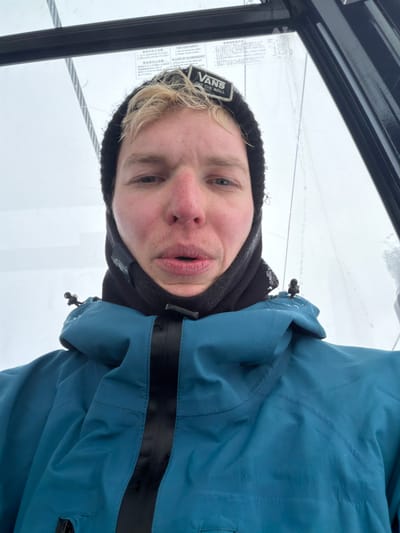 A pale-skinned person with blonde hair wears a black beanie with a Vans logo, a black neck warmer, and a blue jacket with a black stripe across the chest. The person is likely in a gondola, as evidenced by the cable and frame visible in the background. The background appears to be snowy or cloudy. The person looks directly at the camera with a slightly puckered expression.