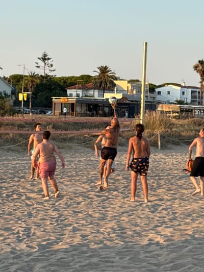 The image captures a group of young men on a sandy beach in Castelldefels, Spain. The sun is low in the sky, casting long shadows and suggesting it is either early morning or late afternoon. The weather appears clear and pleasant.

Several men are visible, most of them shirtless and wearing swim trunks. One man in the foreground is captured mid-action, reaching for a volleyball. Another man stands near him, looking towards the camera. Further back, two other men are walking on the sand, one with a distinctive tattoo on his arm. A fifth man, higher up in the frame, is also walking on the beach. The beach itself is wide and sandy, with ripples and footprints visible, indicating recent activity.

In the background, there are glimpses of buildings and vegetation. A white building with a red-tiled roof stands out, possibly a house or a small hotel, along with other structures that appear to be part of a coastal town. Lush green trees, including palm trees, are scattered across the landscape, adding to the tropical or Mediterranean feel of the location.

A tall, thin pole, possibly a lamppost or a marker, runs vertically through the left side of the image. A yellow sign with some indistinct text is visible on the right side of the background, partially obscured by trees. The overall mood of the image is casual and active, with the individuals appearing to be enjoying their time outdoors.