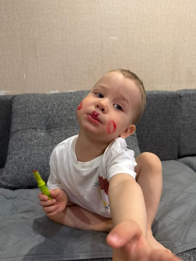 A young child is lying on a gray sofa with lipstick smeared on their face. They are holding a tube of lipstick in their hand. The location is in a residential area. I have no way to determine the city or country from this image.