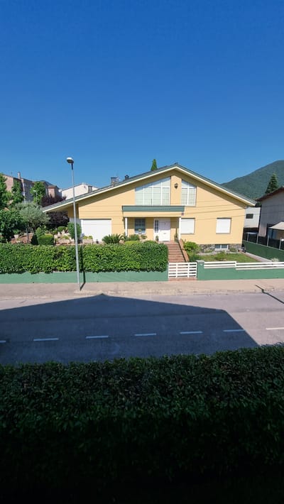 A bright, sunny day is depicted with a clear blue sky above a suburban scene in Sant Joan Les Fonts, Spain. The focal point is a light yellow, two-story house with a large gabled roof. The house features numerous windows, some with white frames and others with shutters. A white garage door is visible on the left side of the house, and a white front door with a glass pane is centrally located on the raised front porch, accessed by a set of stairs with white railings.

The house is surrounded by a well-maintained garden. A thick, dark green hedge runs along the front of the property, creating a boundary with the sidewalk and road. Behind the hedge, there are various plants, including small trees and bushes, and some decorative potted plants. A white picket fence encloses a portion of the front yard.

To the left of the house, a lamppost stands tall near the sidewalk. The road in front of the house has two white dashed lines marking the lanes. A shadow from the lamppost stretches across the road. The background features other houses, trees, and a green, tree-covered hill under the clear blue sky. The overall impression is one of a quiet, well-kept residential area on a beautiful day.