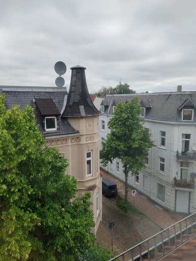 The image captures an elevated view in Neumünster, Germany, overlooking a street scene. Buildings with grey tile roofs and distinct architectural features, including dormers and turrets, line the street. Satellite dishes are visible on one rooftop. Lush green trees partially obscure the buildings. A dark van is parked on the brick-paved street. The sky is overcast. A metal railing is present in the foreground, suggesting the vantage point is from a balcony or window.