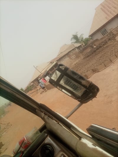 The image depicts a street scene in Oko Erin, Nigeria, captured from inside a vehicle, likely a tricycle, with a significant clockwise tilt. The foreground reveals a portion of the vehicle's dashboard, including a black control knob and a plastic bottle with a red and white label, resting near a worn beige interior trim and the windshield frame.
The midground features an unpaved, reddish-brown dirt road. On the right, a rectangular street light or solar panel unit, framed in black, is mounted on a light-colored pole. Further down the street, on the left side, two to three individuals are partially visible. One person wears a patterned dress (blue, white, black), and another wears a red garment, standing near piles of reddish-brown bricks or concrete blocks stacked along the roadside.
The background consists of multiple single and two-story buildings with light brown walls and corrugated metal roofs, some exhibiting signs of rust. Several buildings feature small, rectangular windows with white frames. A cluster of green palm fronds is visible behind some structures. The sky is hazy and light, suggesting an overcast day or atmospheric dust. No clear actions or direct interactions are discernible among the distant figures.
